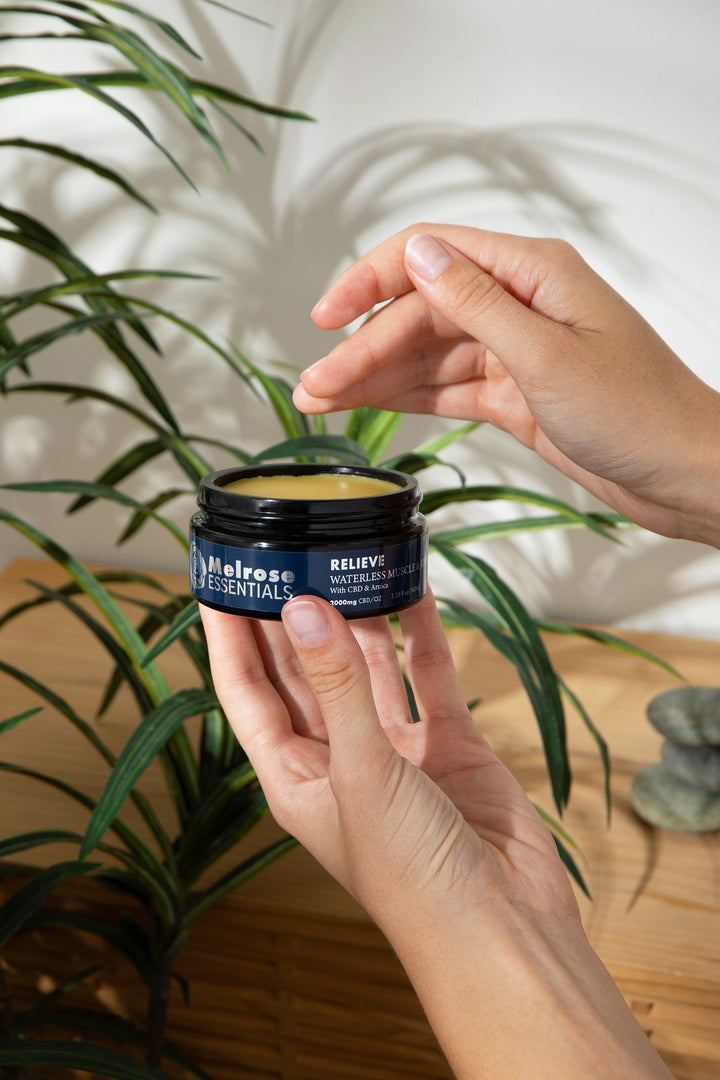 An open jar of Melrose Relieve waterless CBD rub held in two hands beside spa stones